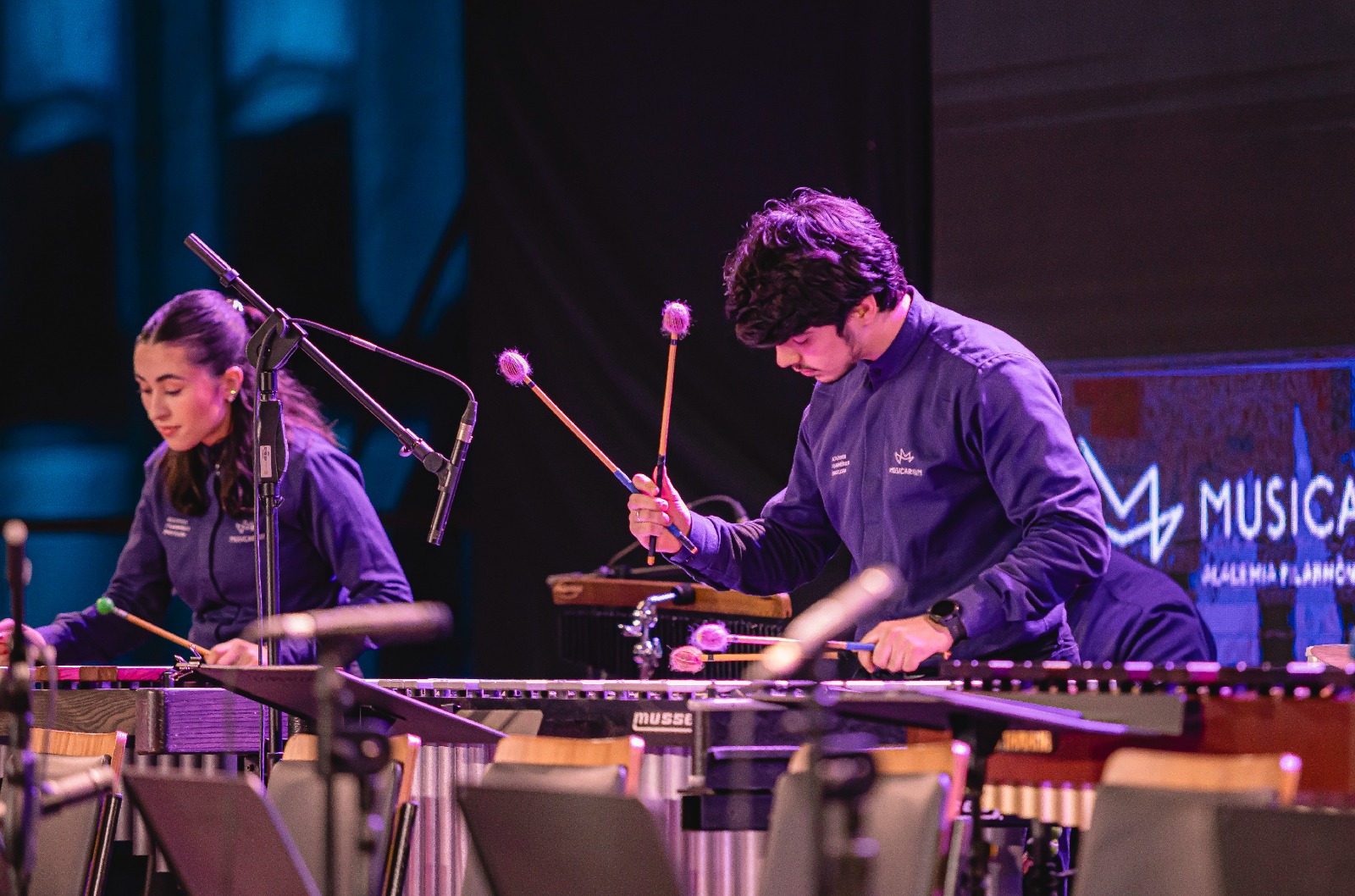 Foto na horizontal mostra uma mulher e um homem jovens com camisas sociais e a logo do Musicarium tocando instrumentos em um palco com iluminação cênica.