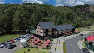 Foto horizontal aérea mostra um casarão bege em estilo enxaimel. À frente dela, algumas mesas ao ar livre com guarda-sóis . Ao lado, uma calçada larga por onde caminham várias pessoas. Ao fundo do casarão, um cinturão verde.