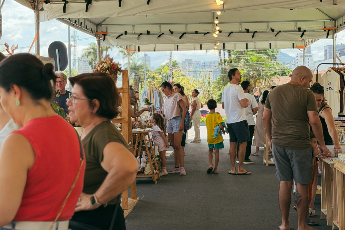 Foto horizontal mostra várias pessoas observando produtos expostos no Festival Meraki, na Praça Cidade das Águas. Os expositores estão debaixo de uma grande estrutura metálica com lona branca. Ao fundo, aparecem algumas árvores e prédios.