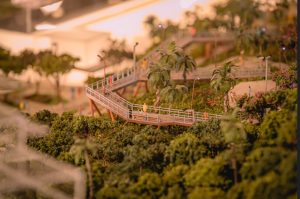Foto horizontal mostra parte de uma maquete onde se vê muitas árvores e uma grande passarela de madeira, com parapeito de metal passando por entre elas. Há também bonecos que simulam pessoas. Ao redor da passarela há postes de iluminação.