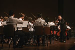 Foto horizontal mostra dezenas de músicos jovens do Musicarium sentados em cadeiras de madeira tocando instrumentos musicais. Eles estão em cima de um grande palco de madeira e de costas para a foto. À frente deles, o maestro Sergio Ogawa. Ele é um homem de meia-idade, média estatura, magro e com traços fisionômicos orientais.