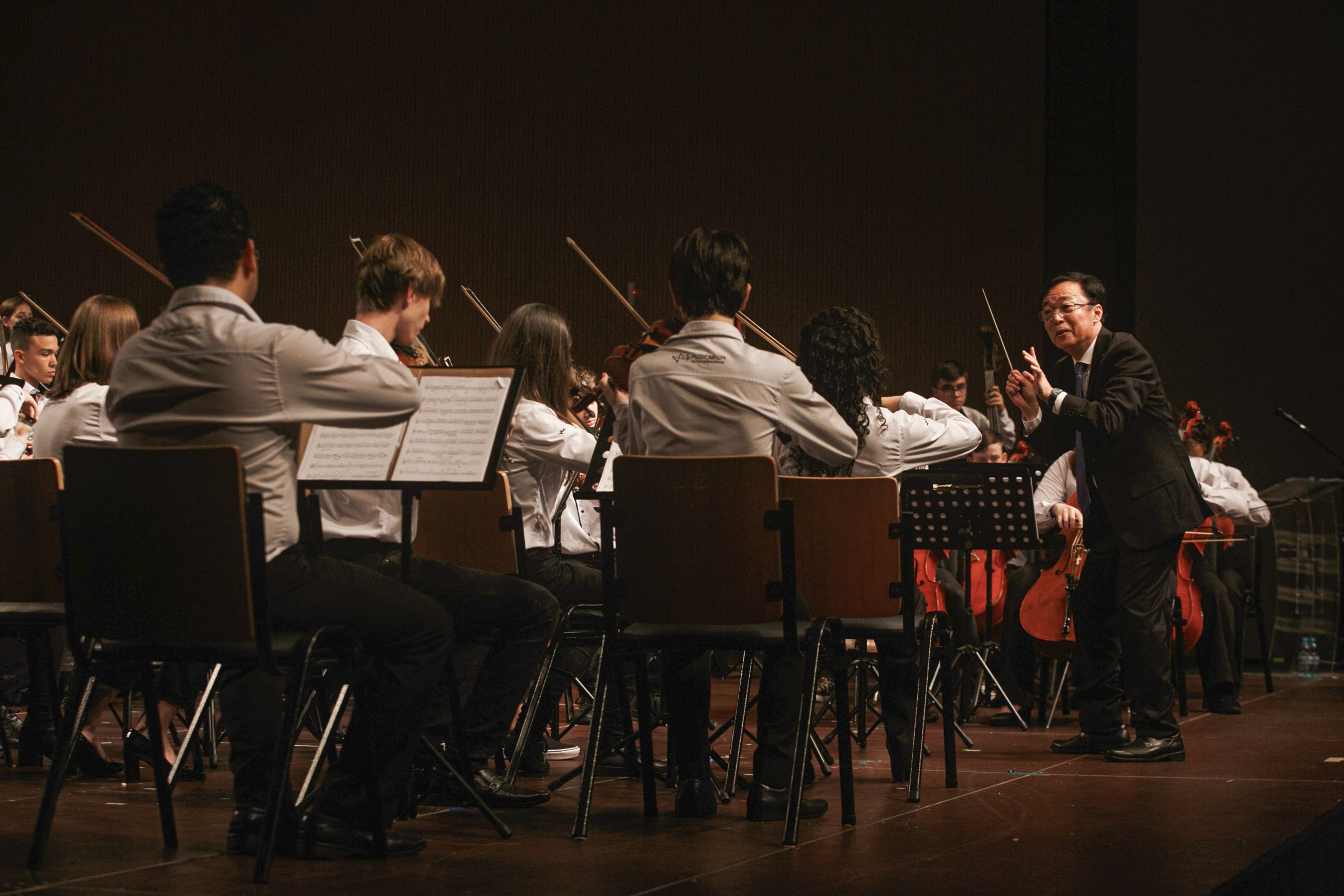 Foto horizontal mostra dezenas de músicos jovens do Musicarium sentados em cadeiras de madeira tocando instrumentos musicais. Eles estão em cima de um grande palco de madeira e de costas para a foto. À frente deles, o maestro Sergio Ogawa. Ele é um homem de meia-idade, média estatura, magro e com traços fisionômicos orientais.