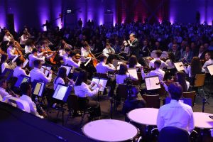 Foto horizontal mostra o maestro do Musicarium Sergio Ogawa regendo uma orquestra de músicos jovens. Sérgio é é um homem de meia-idade, pele parda, cabelos curtos e pretos, média estatura e com traços fisionômicos orientais. Ele usa terno e calça pretos. Atrás dele dezenas de pessoas em um grande salão onde ocorrem as palestras principais da ExpoGestão.