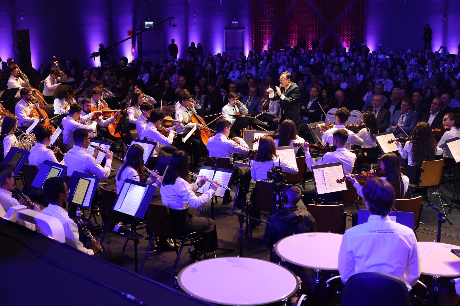 Foto horizontal mostra o maestro do Musicarium Sergio Ogawa regendo uma orquestra de músicos jovens. Sérgio é é um homem de meia-idade, pele parda, cabelos curtos e pretos, média estatura e com traços fisionômicos orientais. Ele usa terno e calça pretos. Atrás dele dezenas de pessoas em um grande salão onde ocorrem as palestras principais da ExpoGestão.