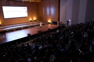 Foto horizontal mostra o grande auditório da Coree Internacional School. Sobre o palco está uma mulher adulta falando ao microfone, durante o Seminário de Educação Inovadora. Atrás dela, um grande telão. De frente para ela e de costas para foto uma plateia lotada de pessoas.
