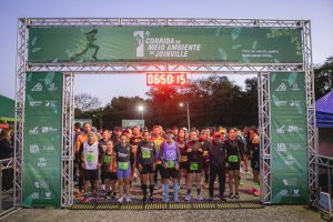 Foto horizontal mostra dezenas de pessoas embaixo de uma grande estrutura metálica com um lona verde da 1ª Corrida do Meio, que é a largada da corrida.