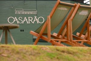 Foto horizontal mostra parede de madeira pintada de verde com a logomarca do Casarão. À frente dela, desfocada, há duas espreguiçadeiras e uma pequena mesa de apoio em uma área gramada.
