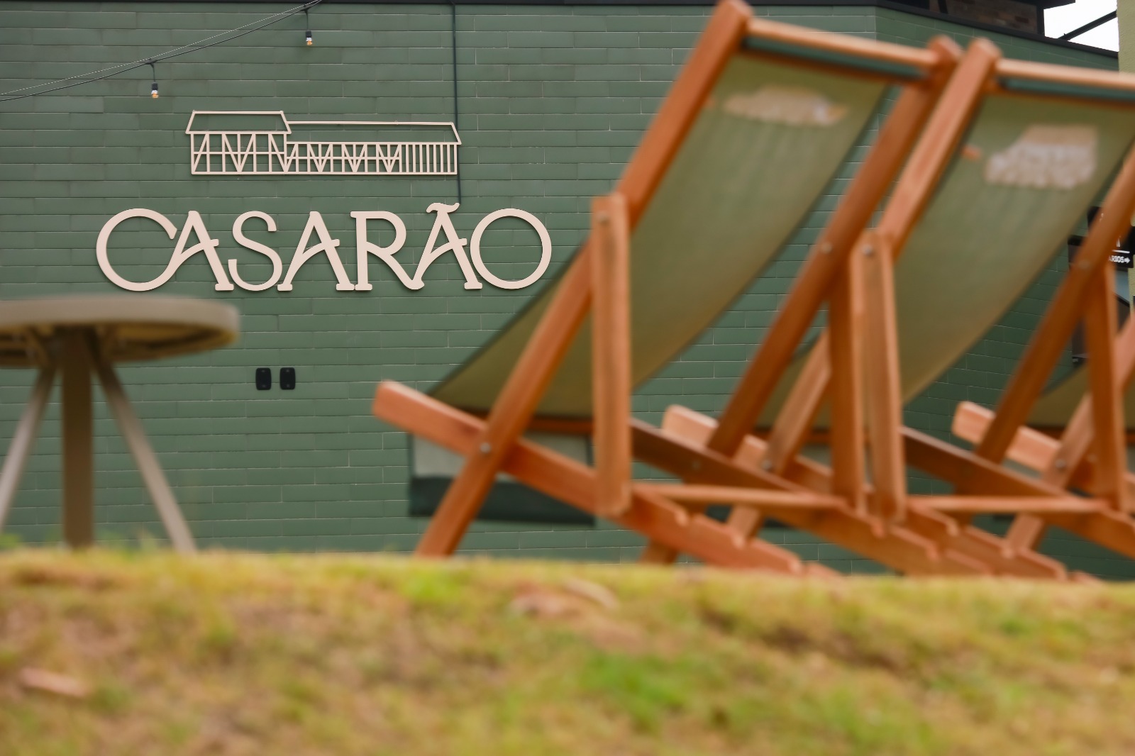 Foto horizontal mostra parede de madeira pintada de verde com a logomarca do Casarão. À frente dela, desfocada, há duas espreguiçadeiras e uma pequena mesa de apoio em uma área gramada.