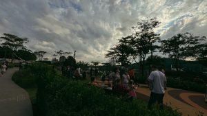 Foto horizontal mostra a praça Cidade das Águas, um amplo espaço gramado, com vários arbustos, árvores e bancos para sentar. Na praça estão dezenas de pessoas. Ao fundo, aparecem algumas bandeiras de festa junina penduradas. O céu está coberto por nuvens e parcialmente nublado.