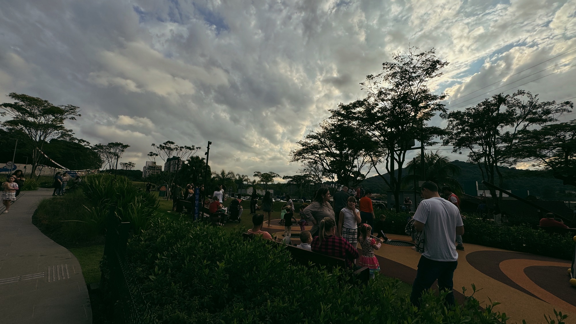 Foto horizontal mostra a praça Cidade das Águas, um amplo espaço gramado, com vários arbustos, árvores e bancos para sentar. Na praça estão dezenas de pessoas. Ao fundo, aparecem algumas bandeiras de festa junina penduradas. O céu está coberto por nuvens e parcialmente nublado.