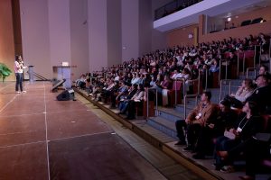 Foto horizontal mostra uma perspectiva lateral do teatro da Coree International School. À esquerda, uma mulher de meia-idade e de cabelos pretos fala ao microfone em um grande palco de madeira, durante o Simpósio de Educação Inovadora Coree 2026. À direita, centenas de pessoas sentadas em cadeiras estofadas em uma plateia, disposta em estilo arena, com vários degraus.