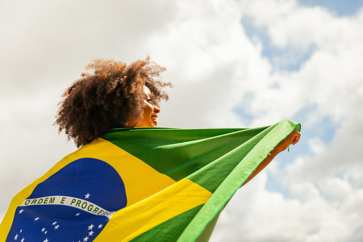 Foto horizontal mostra um mulher jovem, preta, com cabelos castanhos crespos e volumosos em pé, envolvida em uma bandeira do Brasil. Ela está de lado na foto. Ao fundo, o céu azul com nuvens.