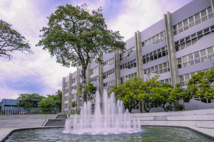 Foto horizontal mostra da fachada do Grupo CRH, um prédio grande, horizontal de cinco andares e paredes cinzas com várias vidraças. Na frente, há um árvore e um grande chafariz ornamental com várias bicas de água jorrando.