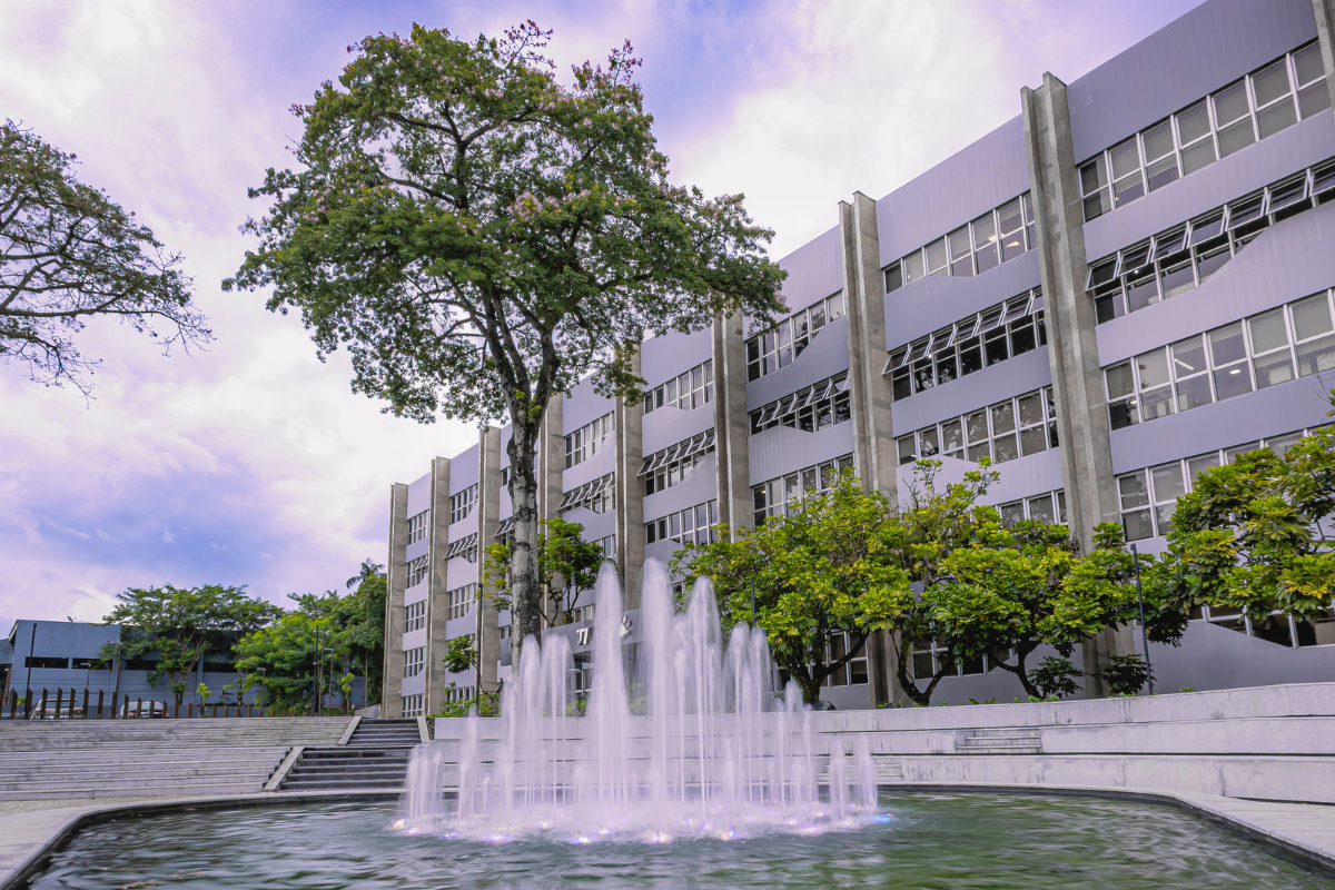 Foto horizontal mostra da fachada do Grupo CRH, um prédio grande, horizontal de cinco andares e paredes cinzas com várias vidraças. Na frente, há um árvore e um grande chafariz ornamental com várias bicas de água jorrando.