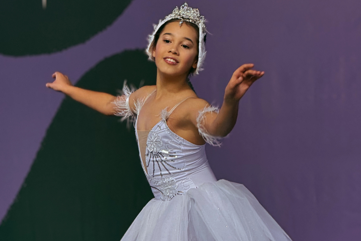 Foto horizontal mostra uma bailarina jovem, de pele parda, olhos castanhos escuros. Ela veste um vestido branco, com uma pequena coroa na cabeça. Ela está com os dois braços abertos e sorri. O fundo é colorido e desfocado.