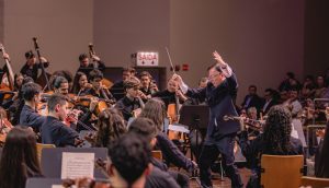Foto na horizontal mostra a orquestra do Musicarium em um grande palco de madeira. Vinte e um jovens, entre homens e mulheres, aparecem na imagem vestindo roupas sociais pretas e estão sentados em cadeiras tocando diferentes instrumentos musicais. À frente deles está o maestro Sérgio Ogawa. Ele é um homem de baixa estatura, pele parda, cabelos curtos e pretos e traços fisionômicos orientais. Ele veste calça social e terno e está com os dois braços erguidos para cima. Na mão direita ele segura uma batuta, pequeno instrumento que rege a orquestra. Do lado direito da foto aparecem parcialmente algumas pessoas na plateia.