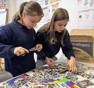 Foto horizontal mostra duas estudantes em frente a uma mesa cheia de figurinhas da Copa do Mundo. Elas são jovens, de pele clara, cabelos lisos e loiros e usam uniforme azul marinho com a logomarca da Coree International School. Elas estão de cabeça baixa mexendo com as figurinhas.