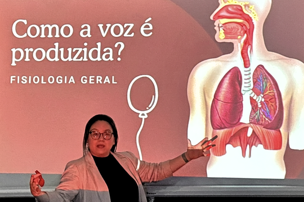 Foto horizontal mostra Idineide Viana Sakata, integrante do Atmen Chor, em frente a uma projeção em um telão. Ela é uma mulher jovem, de pele clara, cabelos lisos e pretos, rosto redondo e olhos levemente puxados, com traços orientais. Ela gesticula com as duas mãos. A apresentação projetada tem como título: "Como a voz é produzida? Fisiologia geral" e mostra o desenho do aparelho fonador humano.