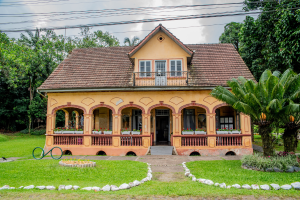 Foto horizontal mostra a fachada da Casa Krüger, um casarão com uma grande varanda, com seis arcos ornamentados, paredes na cor de amarelo envelhecido e balaustres marrons. A casa tem um sótão e telhado estilo duas águas. Na frente da casa há um grande gramado e ao redor aparecem parcialmente algumas árvores.