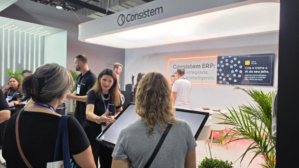 Foto na horizontal mostra o estande da Consistem na Febratex. O espaço tem paredes brancas, mesas e cadeiras claras. Em uma das paredes há um painel com alguns informações, que aparecem parcialmente: "Consistem ERP. Crie e treine a IA do seu jeito". À frente do estande duas mulheres, de costas para foto, conversam com uma mulher de meia-idade e uma blusa preta da Consistem, que gesticula em frente a um totem.