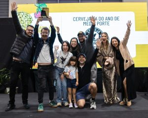 Foto horizontal mostra quatro homens e cinco mulheres, incluindo uma criança, de diferentes idades e características físicas. Eles estão em cima de um pequeo palco, sorriem e estão com as mãos para o alto. Atrás deles, um grande painel, onde aparece parcialmente o escrito: Cervejaria do ano de médio porte e abaixo a logomarca da Stannis, de Jaraguá do Sul.