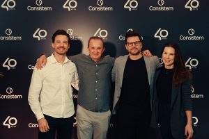 Foto horizontal mostra quatro gestores da Consistem - três homens e uma mulher de pele clara e altos - posando para foto, abraçados, em frente de um grande painel preto com a logomarca da Consistem e o símbolo de 40 anos, em branco. Da esquerda para direita: César Sabel - homem jovem, cabelos curtos castanhos, barba cerrada bom bigode e olhos castanhos; Laureci Sabel - homem idoso, cabelo curto castanho e olhos castanhos; Carlos Sabel - homem jovem, de cabelos curtos castanhos, barba cerrada bom bigode e olhos castanhos, com um óculos retangular; Carla Sabel - mulher jovem, de cabelos longos, lisos e castanhos e olhos castanhos. Todos vestem roupas sociais nos tons de branco, cinza e preto, olham para foto e sorriem.