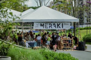 Foto horizontal mostra um conjunto de tendas brancas, debaixo das quais estão vários expositores de diferentes produtos e dezenas de pessoas. À frente da primeira tenda há uma grande faixa branca com a logomarca da Meraki - Feira Colaborativa Multicultural. Ao fundo, aparecem algumas árvores e prédios. No canto inferior da foto, aparecem arbustos da praça Cidade das Águas.