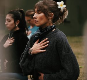 Foto mostra uma mulher jovem, de pele parda, rosto fino e cabelo castanho preso com uma presilha de flor sentada em um gramado com a mão direita no peito e a mão esquerda no umbigo. Ela está de olhos fechados. Ao lado dela, aparecem outras duas mulheres, desfocadas, na mesma posição.
