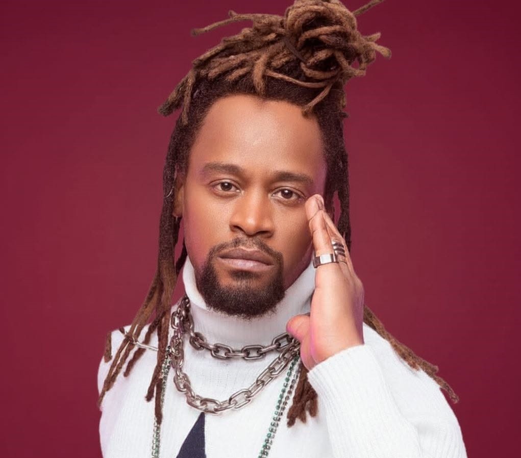 Foto de Toni Garrido. Ele é um homem preto, de meia-idade, rosto alongado, olhos castanhos, lábios grossos, barba e bigode curtos. Seu cabelo tem dreads longos. Ele usa uma blusa de gola alta branca e várias correntes de elo prateadas. O fundo é cor de vinho.