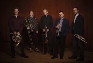 Foto na horizontal de cinco pessoas: quatro homens e uma mulher, integrantes do American Brass Quintet. Todos eles têm pele clara. Eles estão em pé, com roupas pretas, seguram seus instrumentos musicais, olham para foto e sorriem discretamente. Ao fundo, uma parede de madeira.