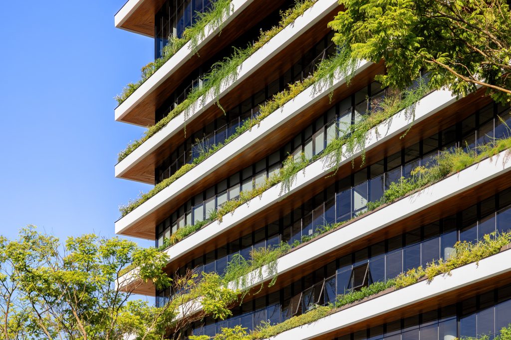 Foto horizontal mostra parte do Edifício Genève, em destaque. O empreendimento é largo, com nove andares e vidraças dispostos de forma diagonal. Em torno de cada andar há uma grande sacada com vegetação.