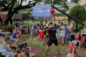 Foto horizontal mostra dezenas de crianças de diferentes idades e características físicas e alguns adultos na Praça Cidade das Águas. Elas estão sobre uma área gramada. Parte delas está em pé, outras correm pelo espaço e algumas estão sentadas. Há adultos com camisetas de recreadores que interagem com elas. No entorno aparecem algumas árvores e uma grande placa azul com a logomarca da Cidade das Águas.